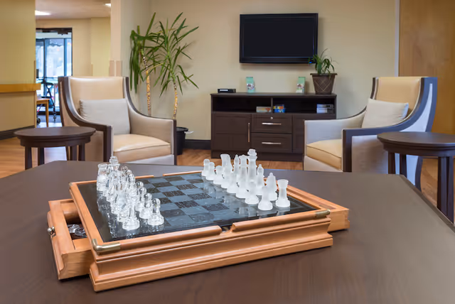 A cozy common area with a wooden table featuring a glass chess set in the foreground. Two beige armchairs with cushions are positioned around the table. In the background, there is a dark wooden cabinet with a flat-screen TV mounted on the wall above it and a potted plant on top. The room has warm lighting and a welcoming atmosphere.