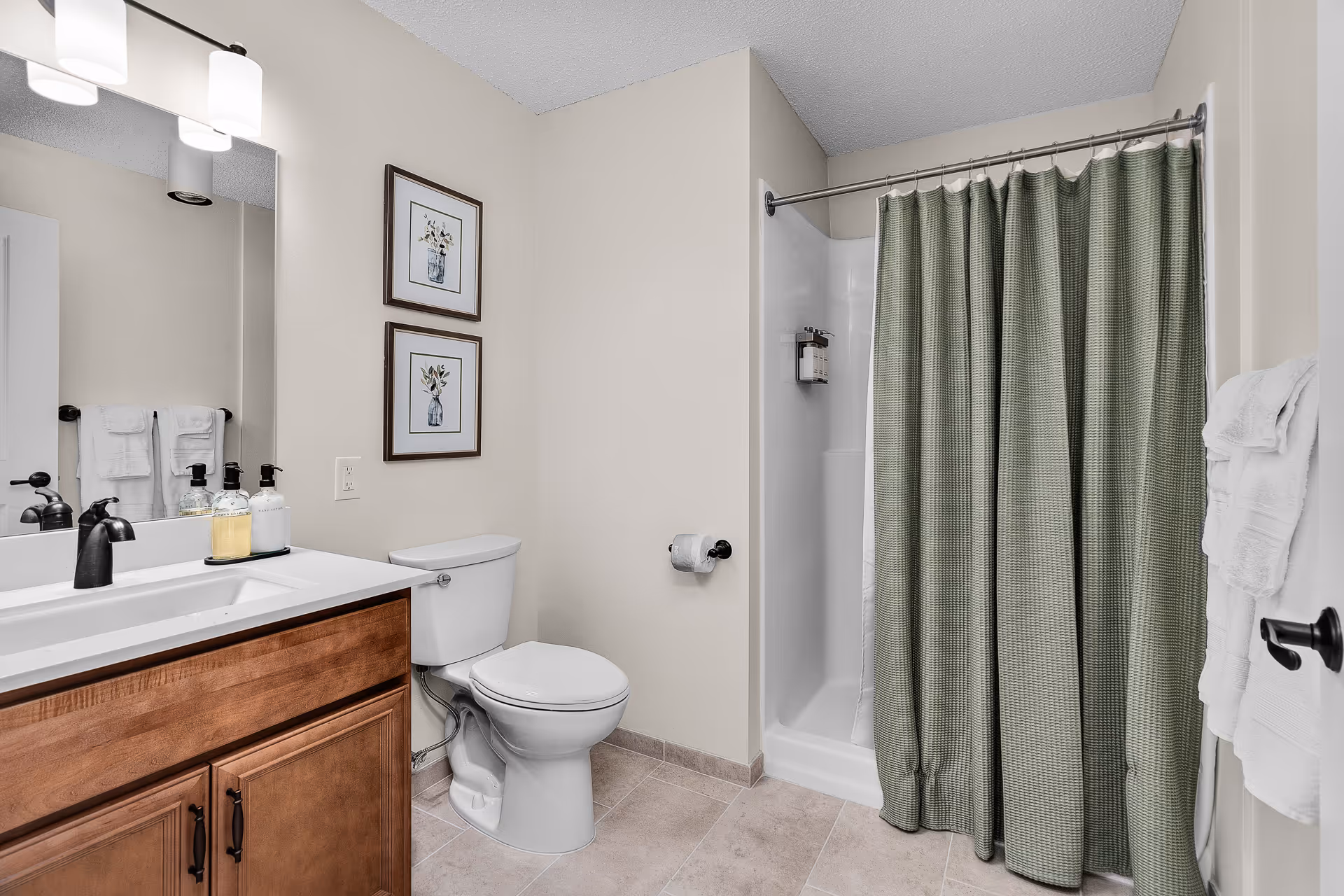 A clean bathroom featuring a wooden vanity with a white countertop and black faucet, a toilet, a shower with a green curtain, two framed floral pictures on the wall, and white towels hanging on racks.