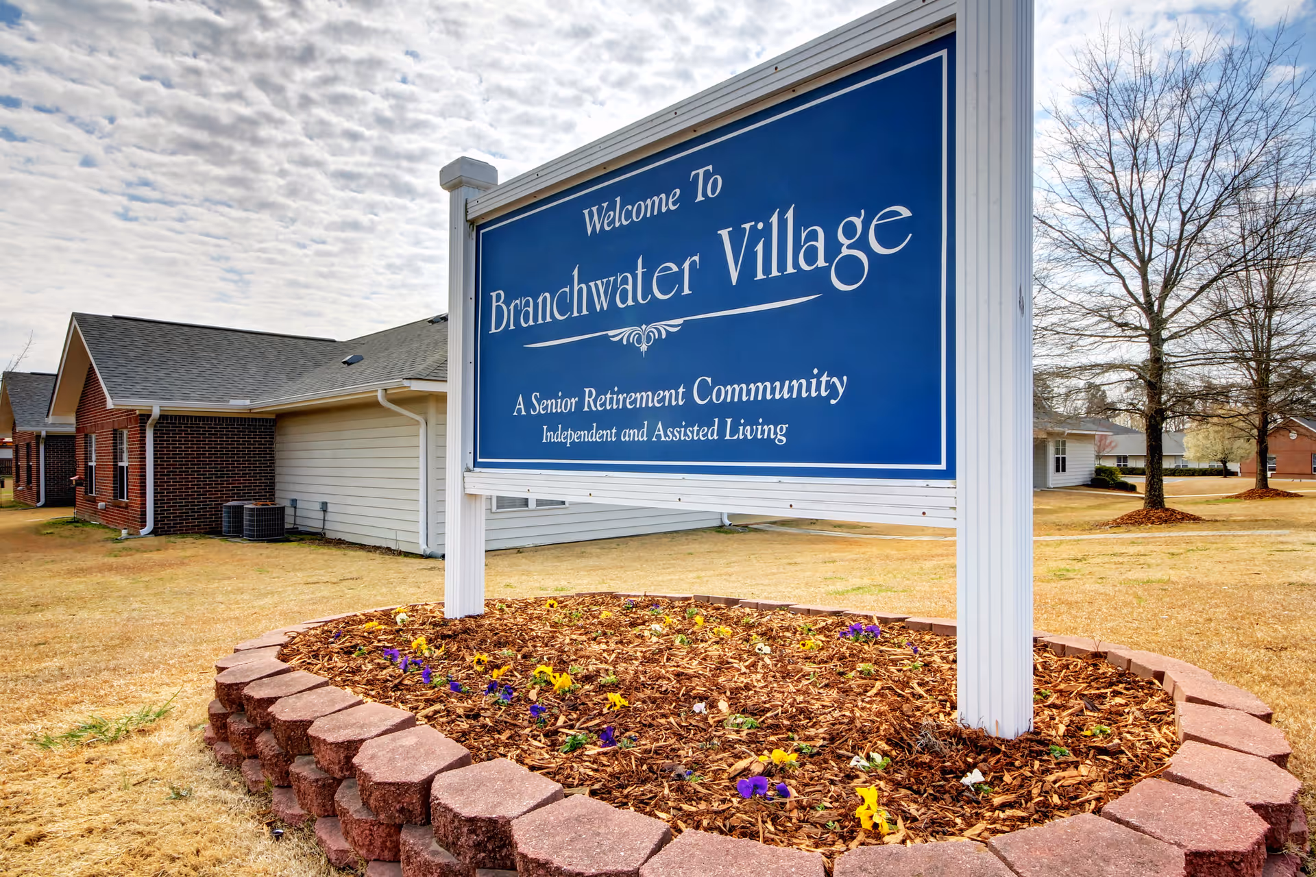A large blue and white sign that reads 'Welcome To Branchwater Village, A Senior Retirement Community, Independent and Assisted Living' is displayed outdoors on a landscaped area with flowers and mulch. In the background, there are single-story brick and siding buildings, a few leafless trees, and a cloudy sky.
