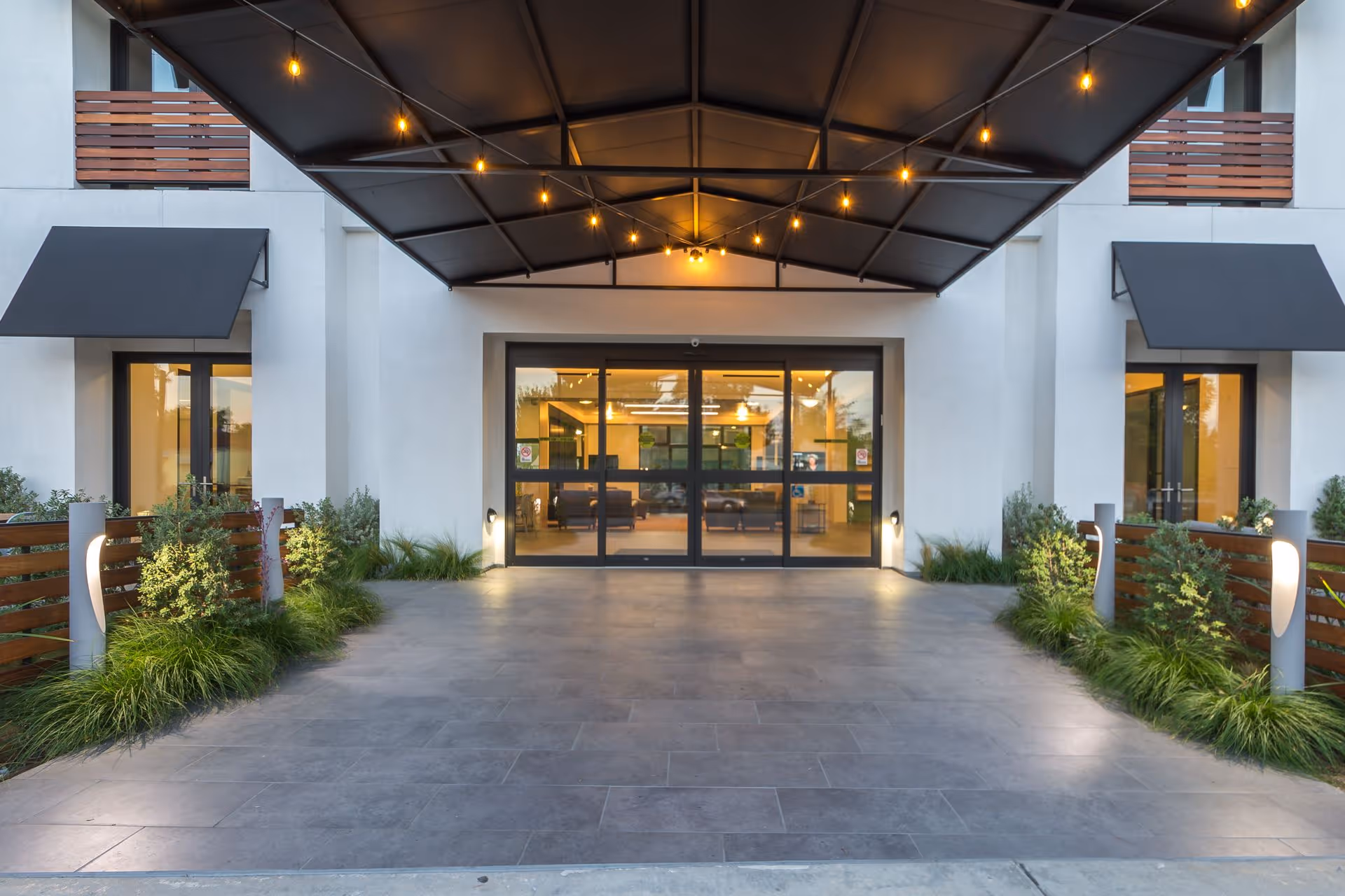 Entrance of a modern senior living facility with a covered driveway, glass automatic sliding doors, and warm lighting inside. There are plants and decorative lights along the sides of the entrance pathway.