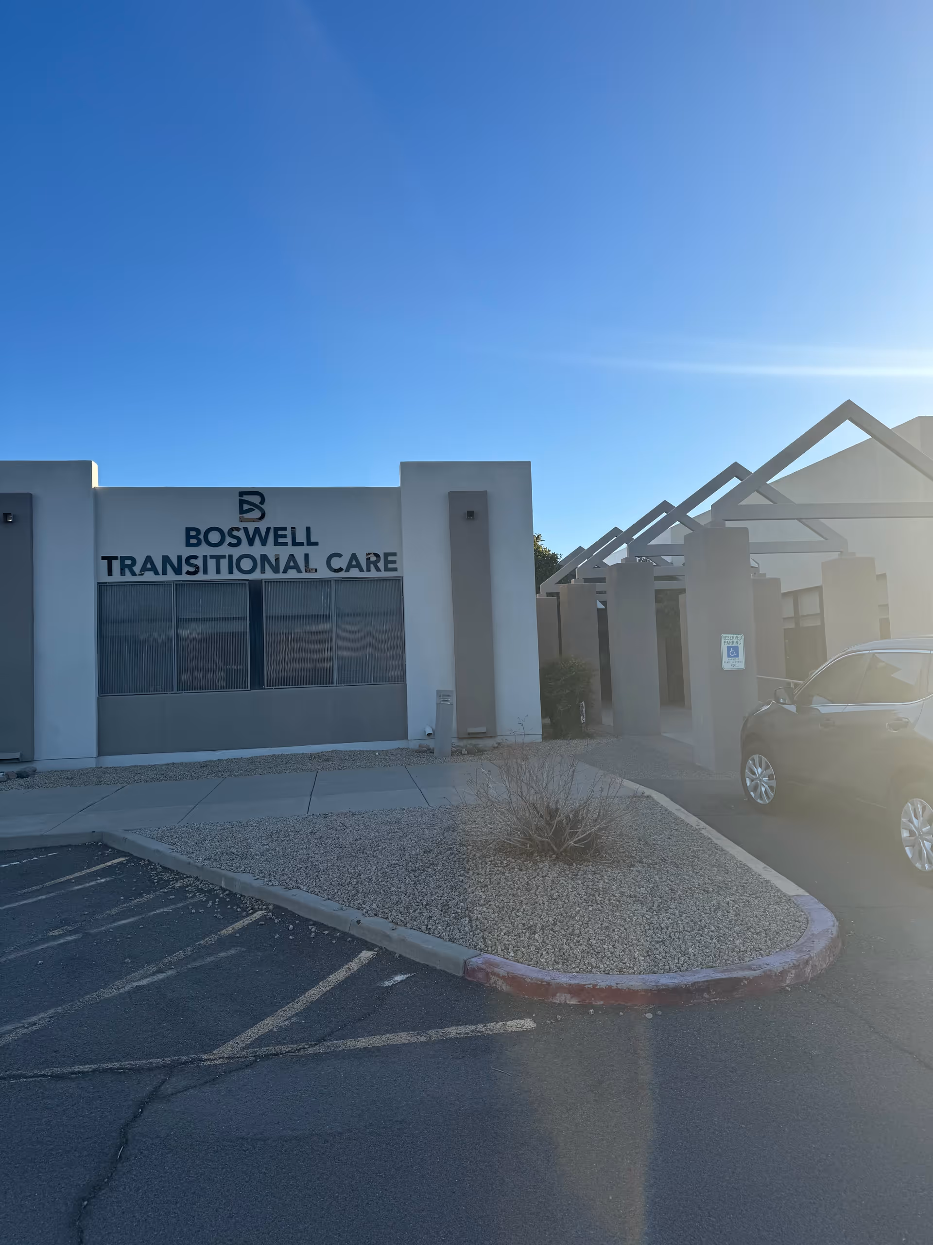 Front exterior of Boswell Transitional Care building with its sign, entrance structure, and a small parking area under a clear blue sky.