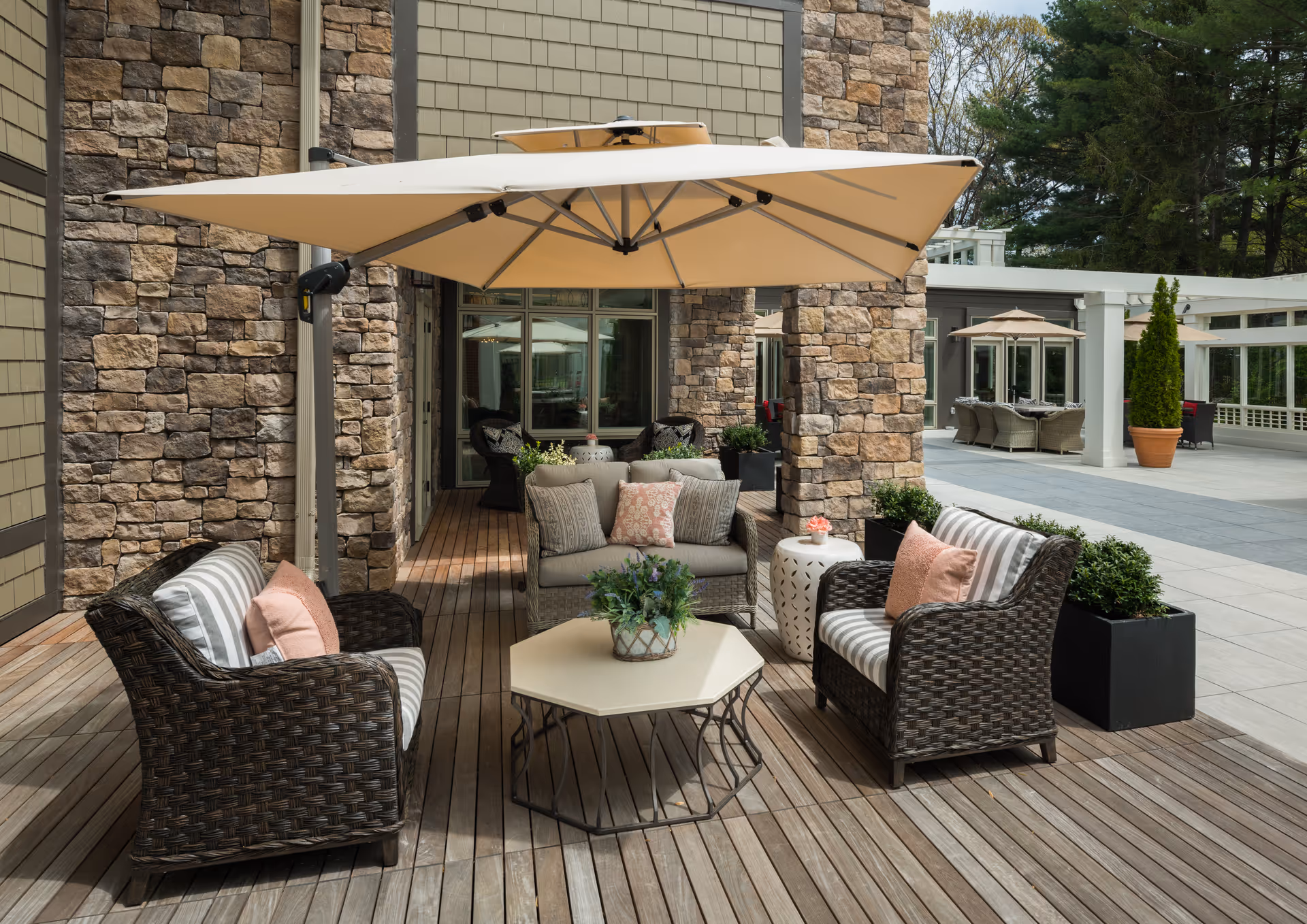Outdoor patio area with wicker furniture including two armchairs and a loveseat with striped and solid cushions, a hexagonal coffee table with a potted plant, large beige umbrella providing shade, stone walls, wooden deck flooring, and additional seating areas in the background surrounded by greenery.