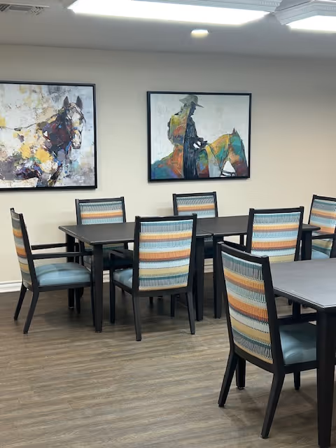 A dining area with several dark wood tables and chairs featuring striped upholstery in shades of blue, orange, and beige. The room has light-colored walls with two framed paintings depicting abstract horse and rider artwork. The floor is covered with wood-patterned flooring.
