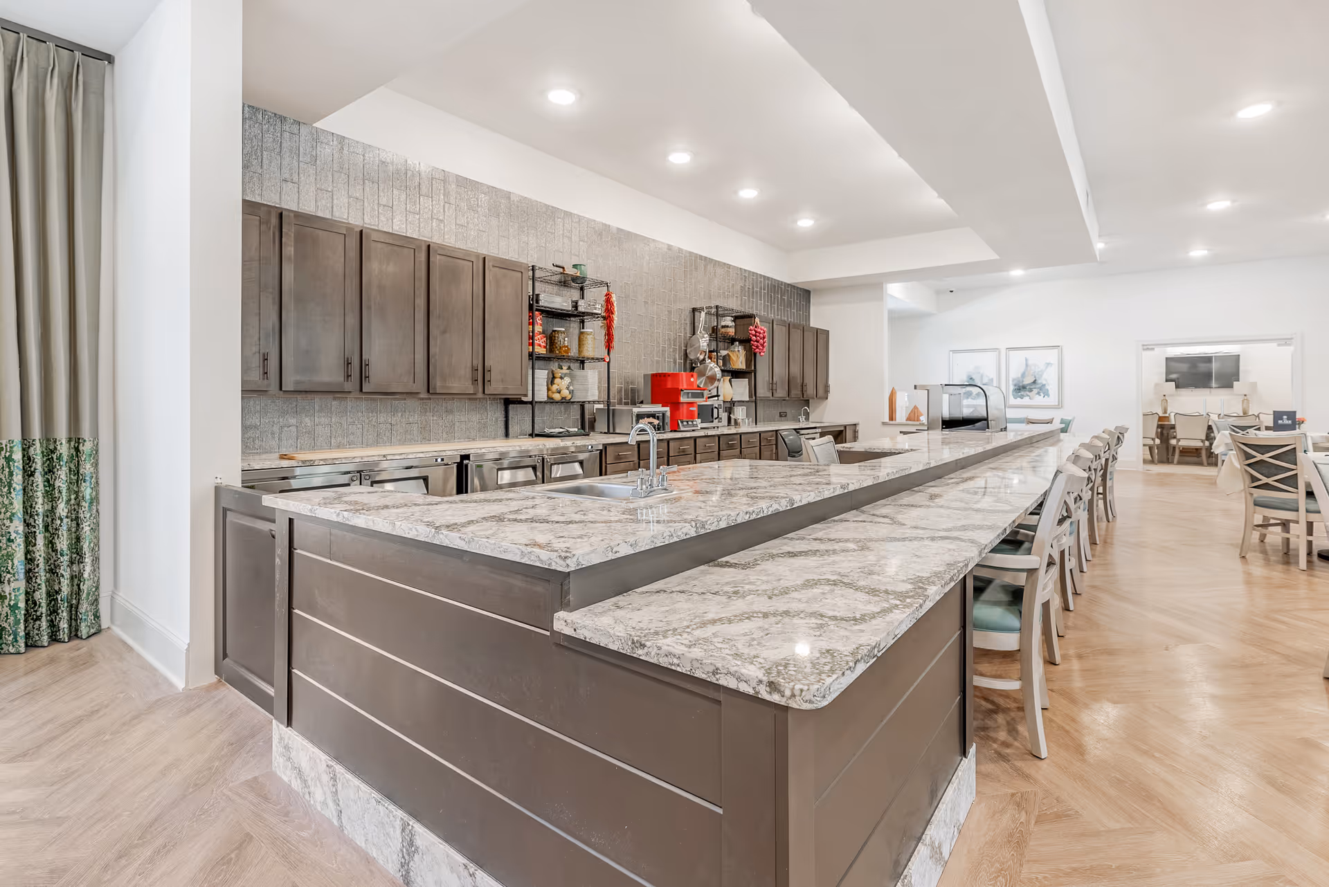 A spacious kitchen area with a long marble countertop island featuring a sink and multiple chairs lined up along one side. The back wall has dark wooden cabinets, open shelves with kitchen items, and a red coffee machine. The room is well-lit with recessed ceiling lights and has light wood flooring. In the background, there is a dining area with tables and chairs.