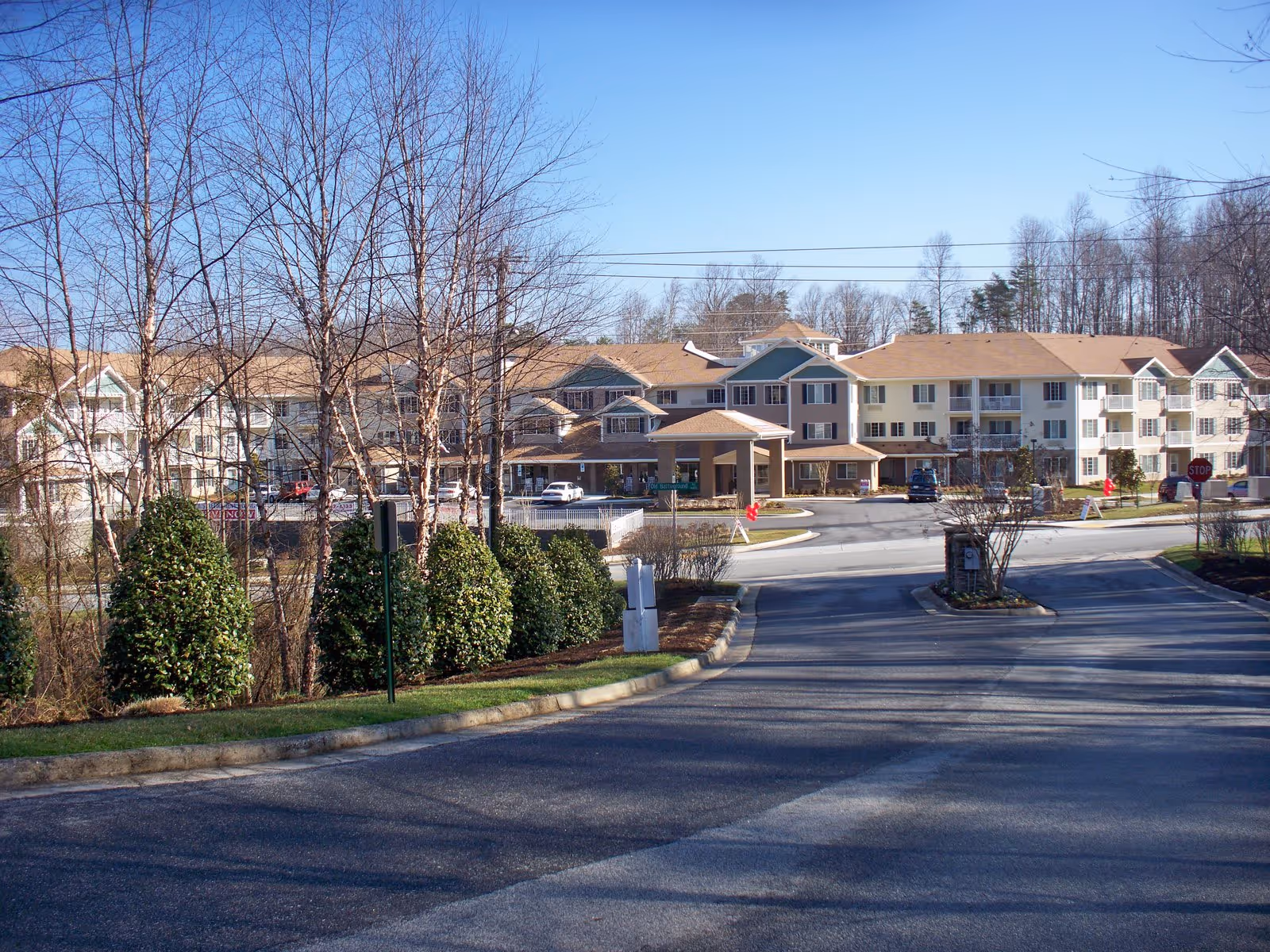 View of the exterior of Carolina Estates Gracious Retirement Living facility on a clear day, showing a multi-story building with beige and green accents, surrounded by trees and bushes, with a paved driveway leading to the entrance.
