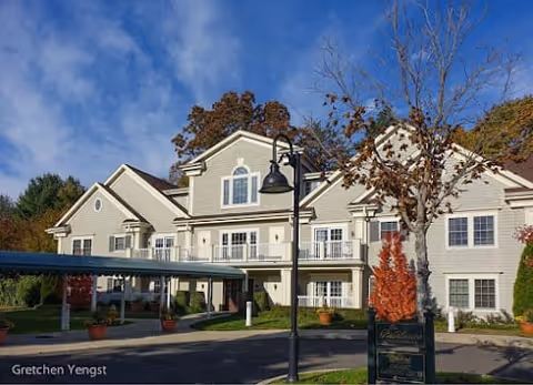 Exterior view of a two-story senior living facility building with beige siding, white trim, and multiple windows. There is a covered driveway entrance with potted plants, a black street lamp, and a tree with autumn-colored leaves in front. The sky is clear and blue.