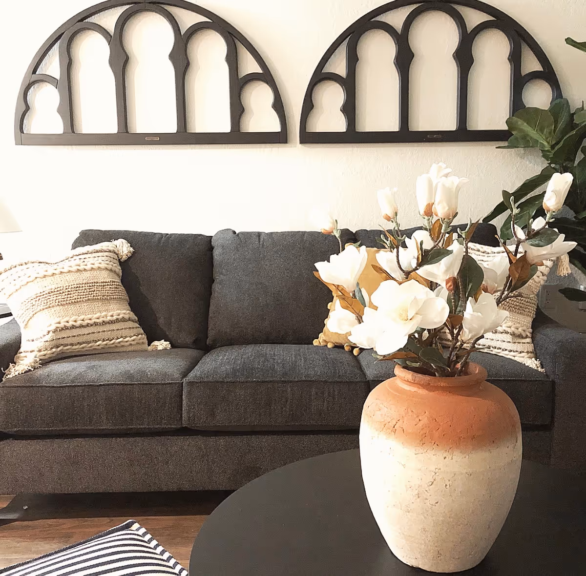 A cozy living room area featuring a dark gray sofa with textured beige and cream pillows. Above the sofa, there are two decorative black wall hangings with arched cutout designs. In the foreground, a round black coffee table holds a large ceramic vase with white flowers. There is a leafy green plant partially visible on the right side.