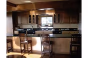 Interior view of a kitchen area with wooden cabinets, a black countertop, and a breakfast bar with four wooden chairs. A window above the sink lets in natural light.
