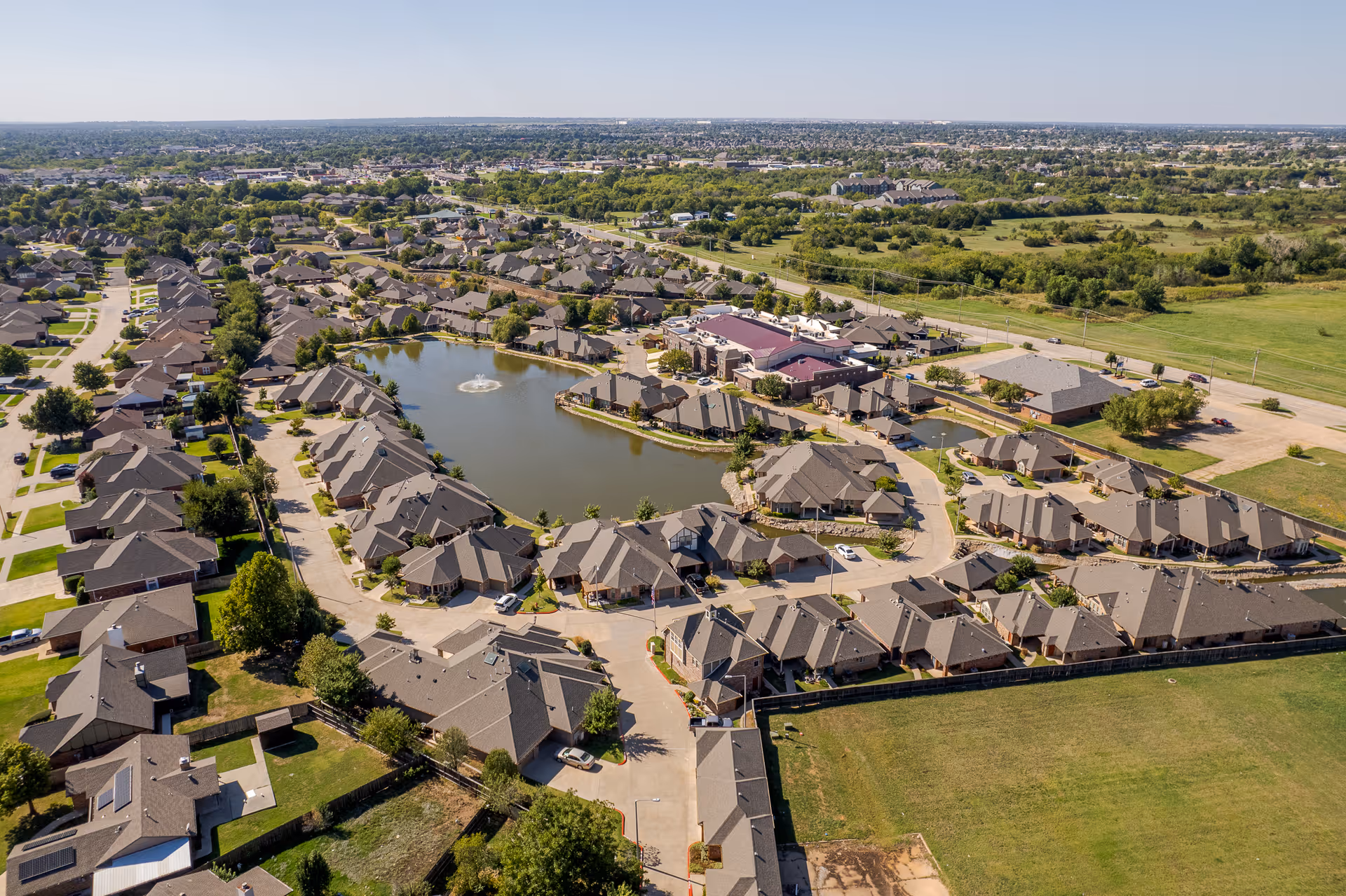 Aerial view of a residential community with multiple single-story buildings arranged around a central pond with a fountain. The area is surrounded by green spaces and roads, with more houses and open land visible in the distance under a clear sky.