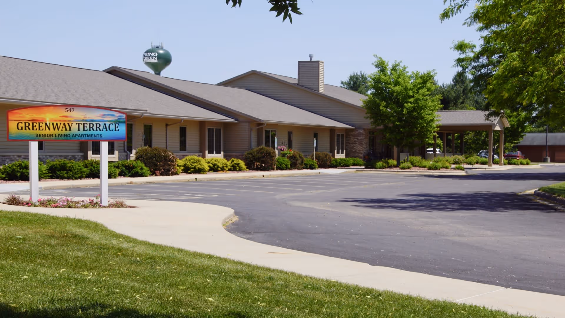 Exterior view of Greenway Terrace Senior Living Apartments building with a parking lot in front, surrounded by green trees and bushes under a clear blue sky.