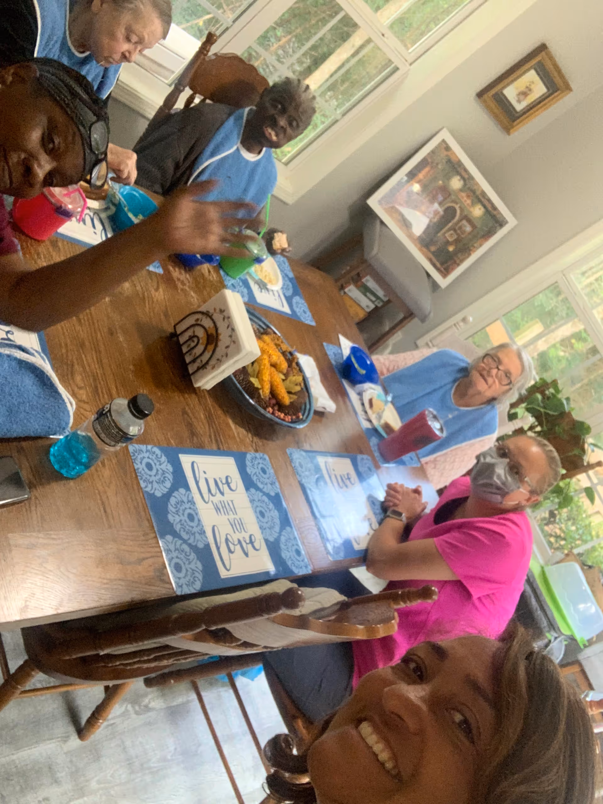 A group of elderly residents and a caregiver seated around a dining table in a bright dining room, sharing a meal and smiling.