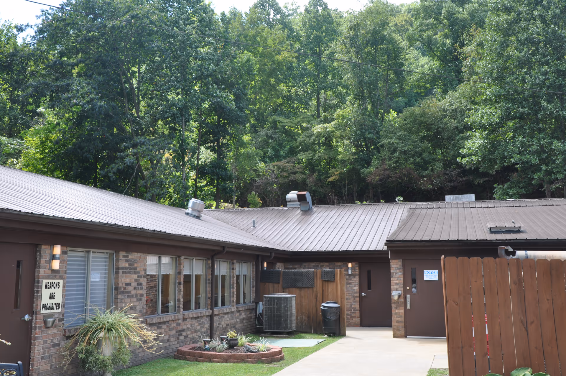 Exterior view of a single-story brick building with a brown metal roof surrounded by tall green trees. There is a small garden area with plants and a sign on the wall that reads 'WEAPONS ARE PROHIBITED'. A wooden fence and a concrete pathway are also visible.