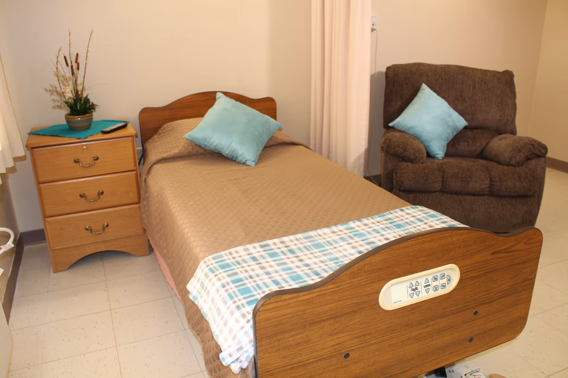 A small bedroom with a single hospital-style bed covered with a brown quilt and a blue plaid blanket at the foot. There is a light blue pillow on the bed. Next to the bed is a wooden nightstand with three drawers, a green plant, and a remote control on top. A brown upholstered armchair with a matching light blue pillow is positioned against the wall. The room has beige walls and a tiled floor.