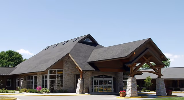 Exterior front view of a senior living facility building with stone pillars supporting a covered entrance, large windows, and a sloped roof under a clear blue sky.