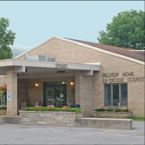 Exterior view of Hillview Home La Crosse County building with a covered entrance, brick walls, windows, and flower beds in front. Trees are visible in the background.