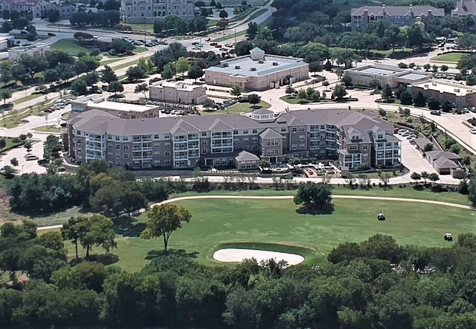 Aerial view of a large multi-story senior living facility named Pioneer Ridge Gracious Retirement Living, surrounded by parking lots, trees, and green spaces including a golf course with sand traps and golf carts visible.