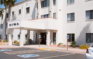 Exterior view of a white multi-story building with several windows and a covered entrance. There are palm trees and landscaped areas with bushes near the entrance. Handicap parking spaces are visible in front of the building.