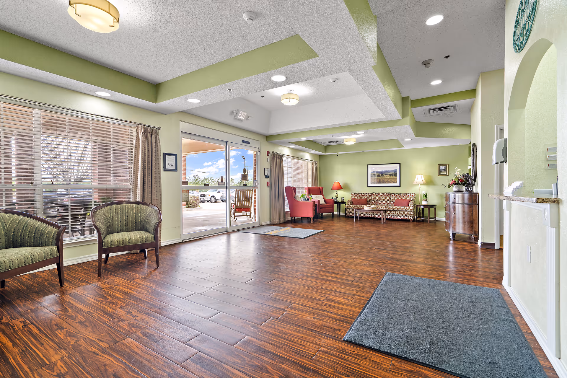 A spacious and well-lit common area with wooden flooring, green walls, and a tray ceiling with recessed lighting. The room features several seating options including two green armchairs near large windows with blinds, two red armchairs, and a patterned sofa with side tables and lamps. There is a glass door leading outside, and decorative elements such as framed pictures and a floral arrangement on a wooden cabinet.