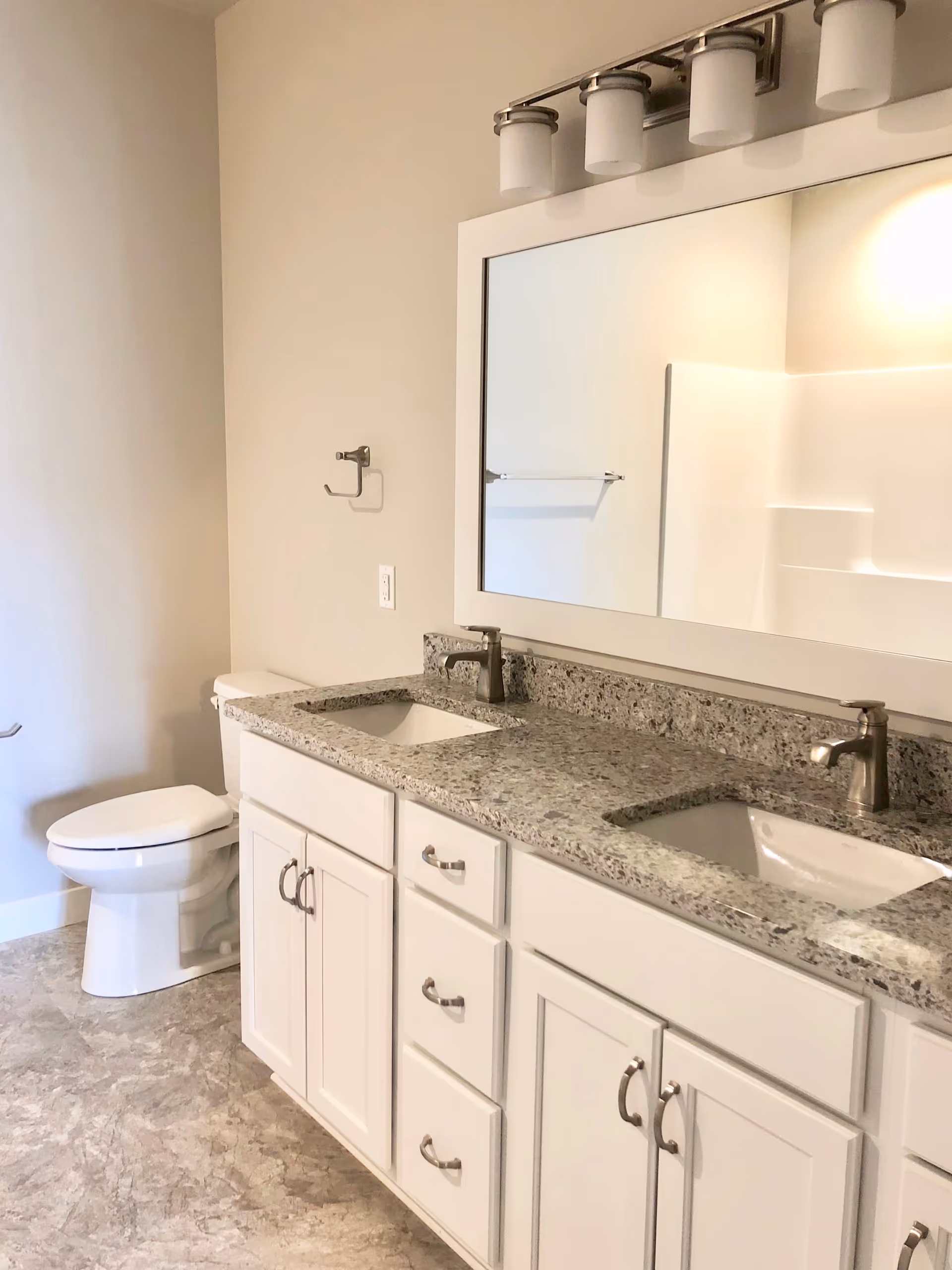 A bathroom with a double sink vanity featuring a granite countertop, white cabinets, and two modern faucets. Above the sinks is a large rectangular mirror with a light fixture containing four lights. To the left of the vanity is a white toilet and a towel ring mounted on the wall. The floor has a stone-like tile pattern.