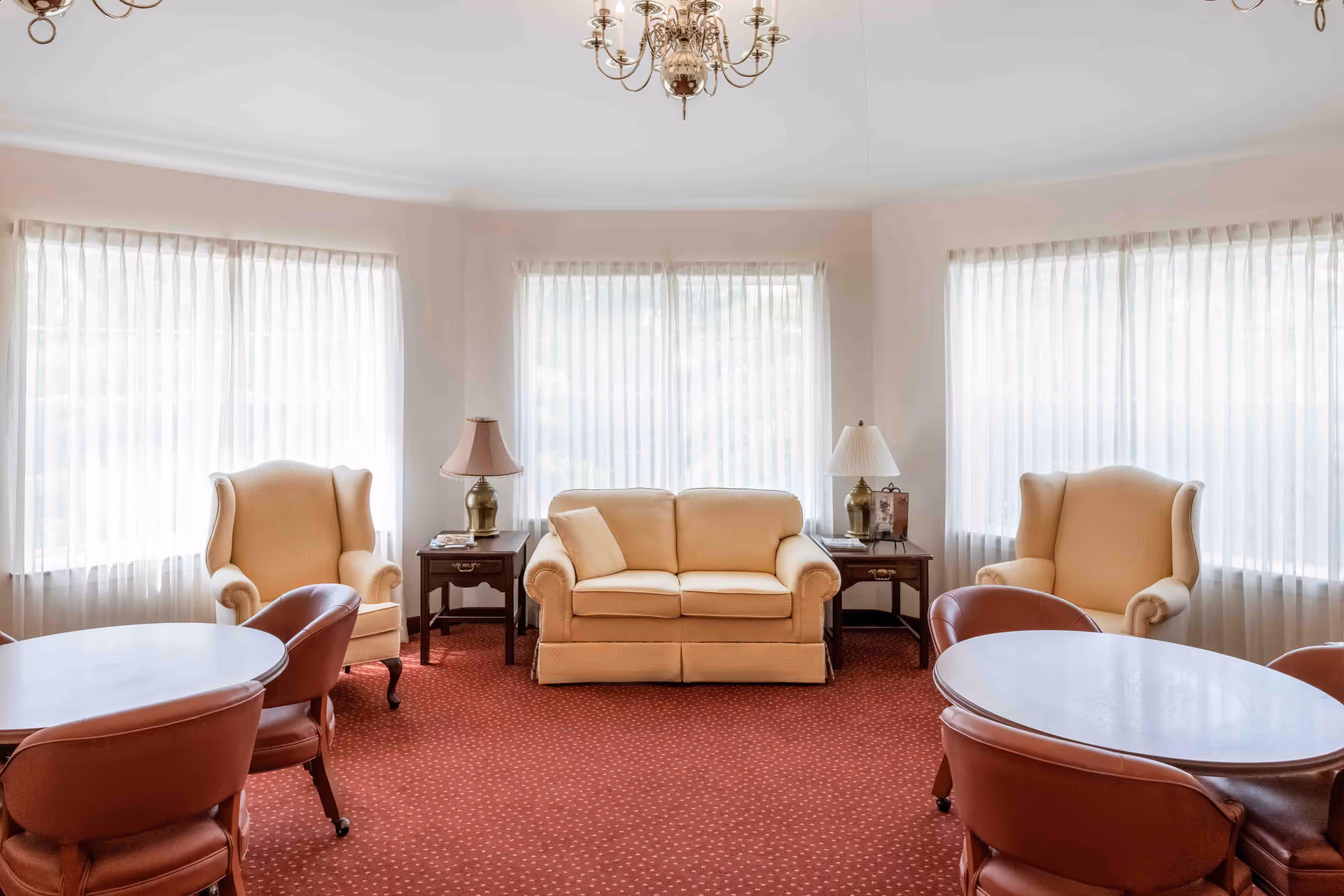 A bright and cozy living room with three large windows covered by sheer white curtains. The room features a beige loveseat with a matching pillow in the center, flanked by two beige wingback chairs. There are two wooden side tables with brass lamps on either side of the loveseat. In the foreground, there are two round tables surrounded by brown leather chairs on casters. The floor is covered with a red carpet with a subtle pattern, and a brass chandelier hangs from the ceiling.