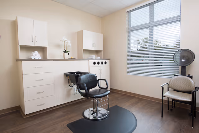 A clean and well-lit hair salon room with a black salon chair in front of a hair washing sink, light wood cabinets with towels and a potted plant on the countertop, a window with blinds letting in natural light, and a beige chair with a hair dryer next to it.