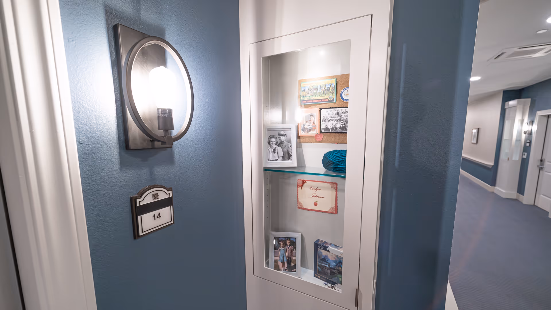 Interior hallway with a blue wall, a lit wall sconce, a room number plaque reading 14, and a glass display cabinet with photos and memorabilia.