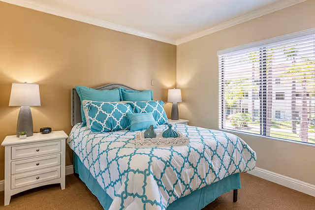 A bright bedroom with a neatly made bed featuring a white and turquoise patterned comforter and multiple pillows. There are two white nightstands on either side of the bed, each with a lamp. A large window with white blinds lets in natural light, showing a view of trees and buildings outside. The walls are painted beige and the room has a clean, cozy atmosphere.