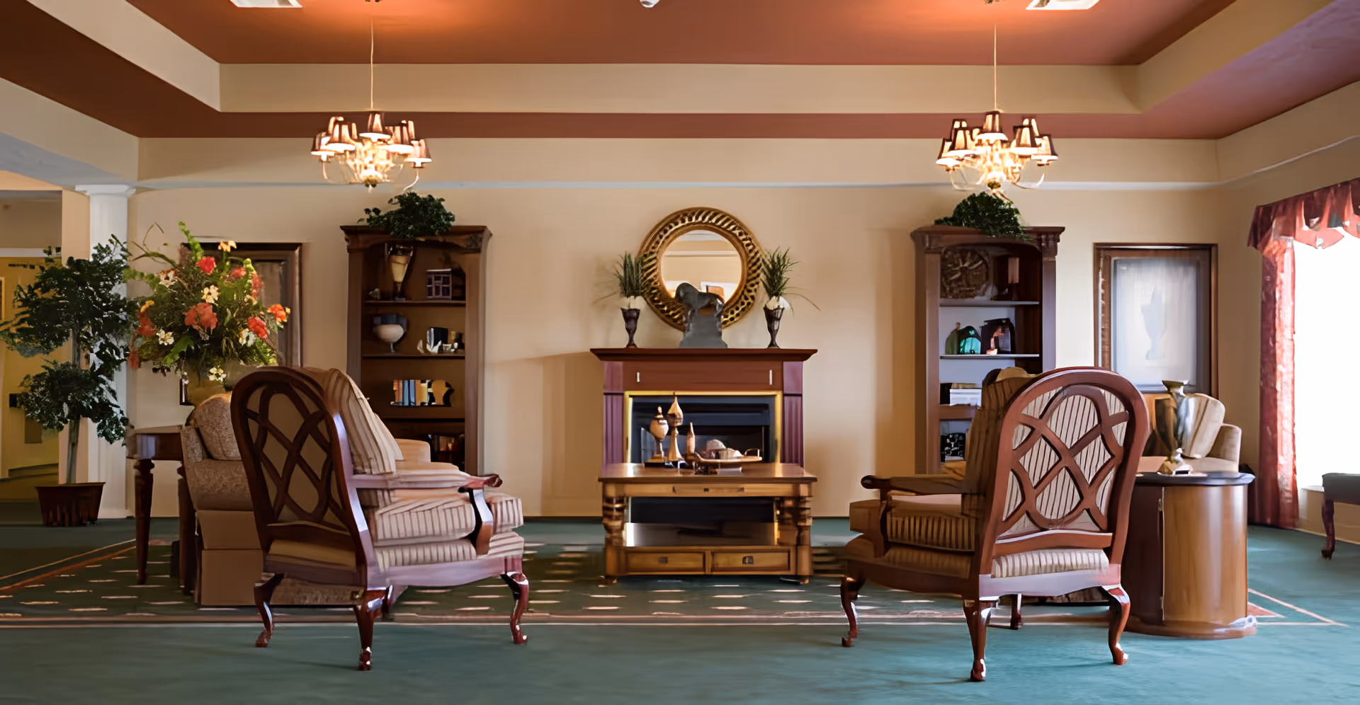 A cozy living room with two wooden armchairs with striped cushions facing a wooden coffee table and a fireplace. The fireplace has a round decorative mirror above it and two vases with plants on either side. There are two wooden bookshelves flanking the fireplace, each decorated with various items. The room has green carpet, beige walls, and a ceiling with recessed lighting and two chandeliers. A large floral arrangement and a potted plant are visible on the left side, and a window with red curtains is on the right.