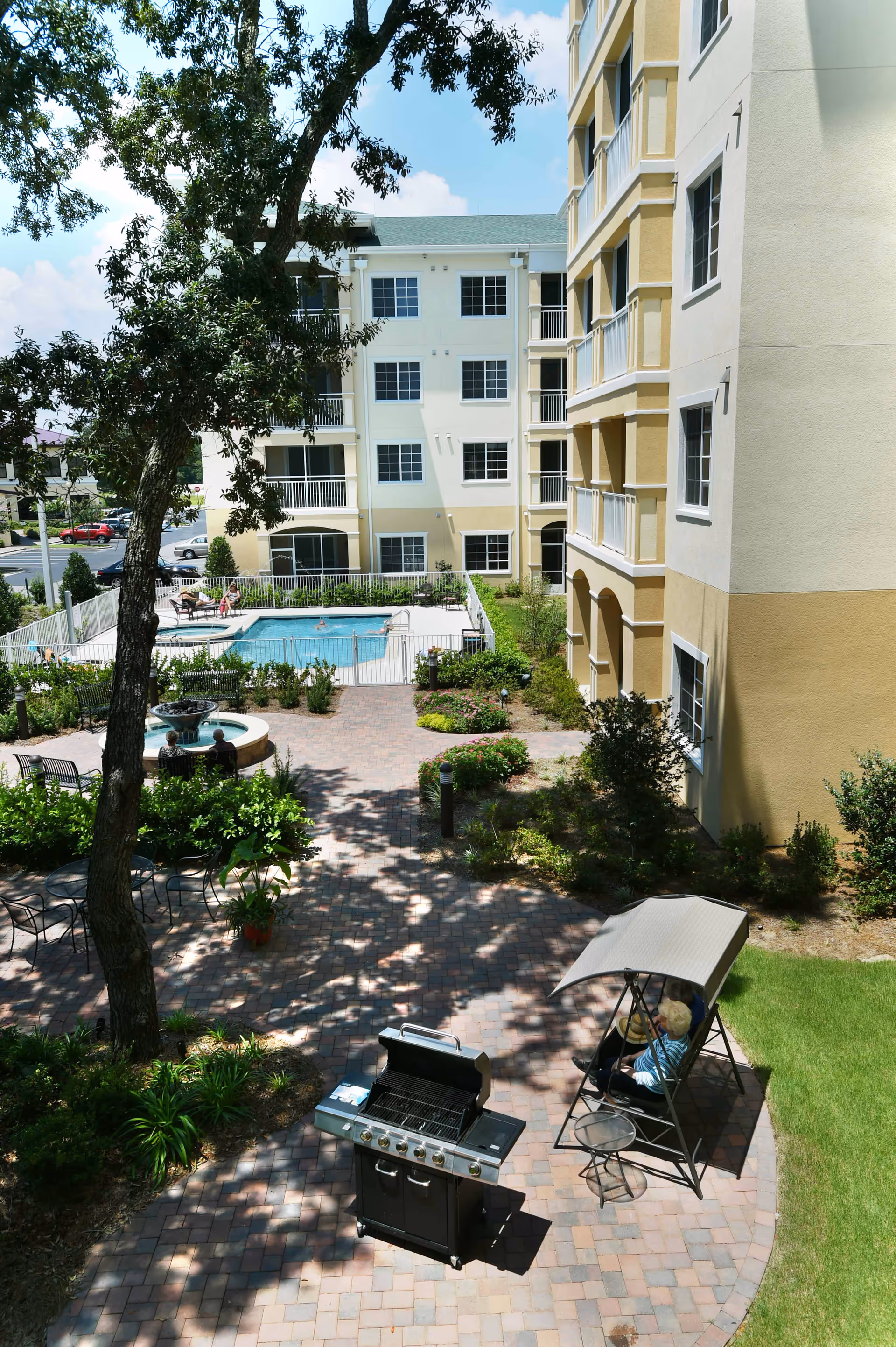 Outdoor courtyard area of a senior living facility with a paved walkway, a barbecue grill, a shaded swing with a person sitting on it, a fountain, and a fenced swimming pool surrounded by lounge chairs. The courtyard is landscaped with trees, bushes, and flowers, and is adjacent to a multi-story building with balconies and windows.