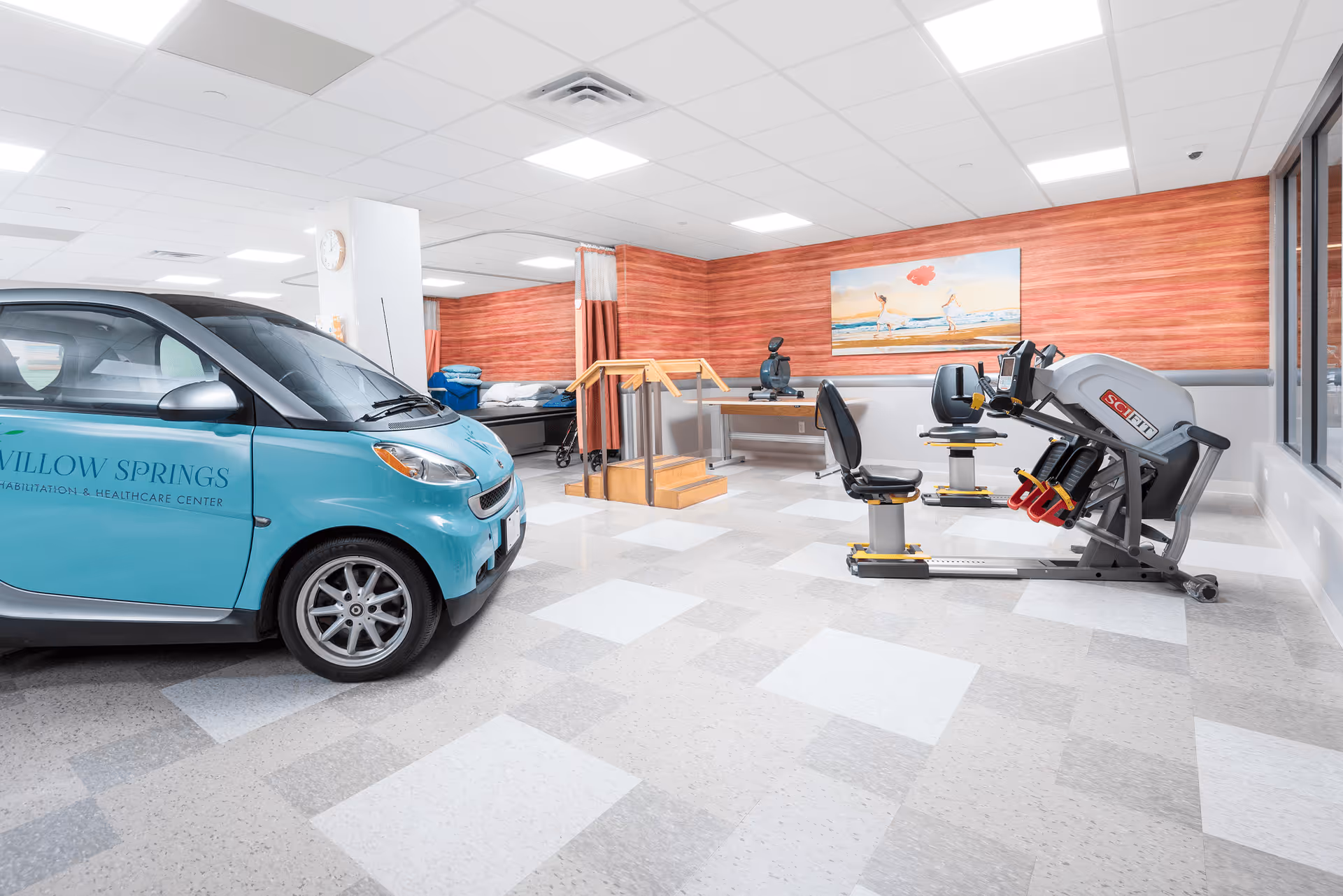 Interior view of a rehabilitation and healthcare center room featuring a small blue car labeled 'Willow Springs Rehabilitation & Healthcare Center', exercise equipment including a seated leg press machine, a stationary bike, a set of stairs with handrails for physical therapy, and a bed with pillows in the background. The room has a tiled floor, wood-paneled walls, and a painting of two people on a beach.