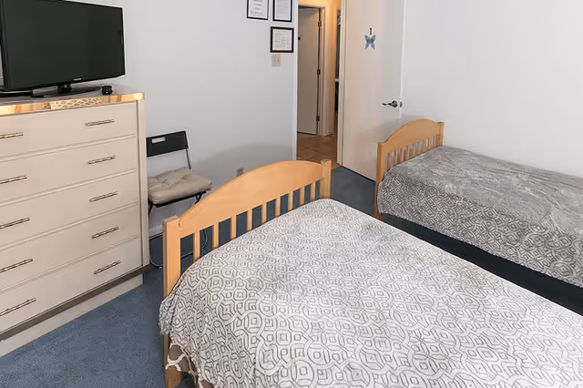 Two twin beds with patterned gray bedspreads in a simple bedroom with a dresser, TV, and an open door to a hallway.