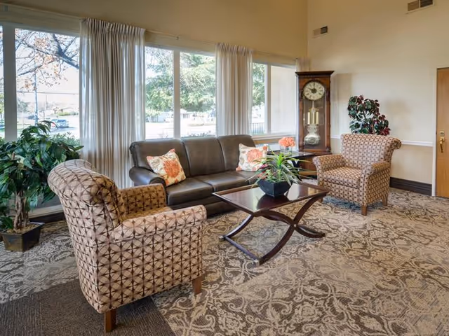 A cozy living room area with a dark brown leather sofa adorned with floral pillows, two patterned armchairs, a wooden coffee table with a plant centerpiece, a grandfather clock, and large windows with white curtains letting in natural light. There are also two potted plants in the corners of the room.