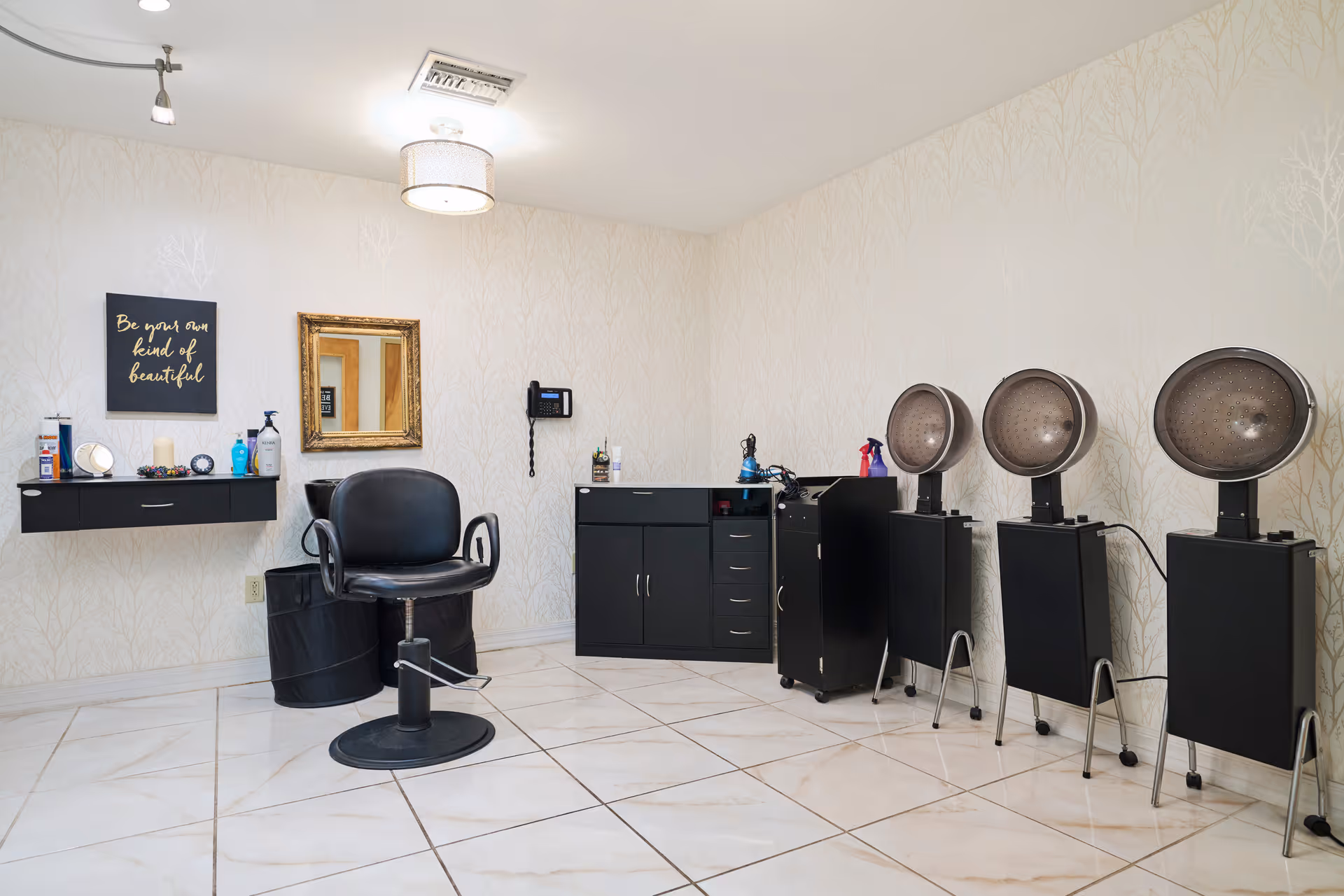 A small salon room with a styling chair, cabinets, three hooded hair dryers and wall decor.