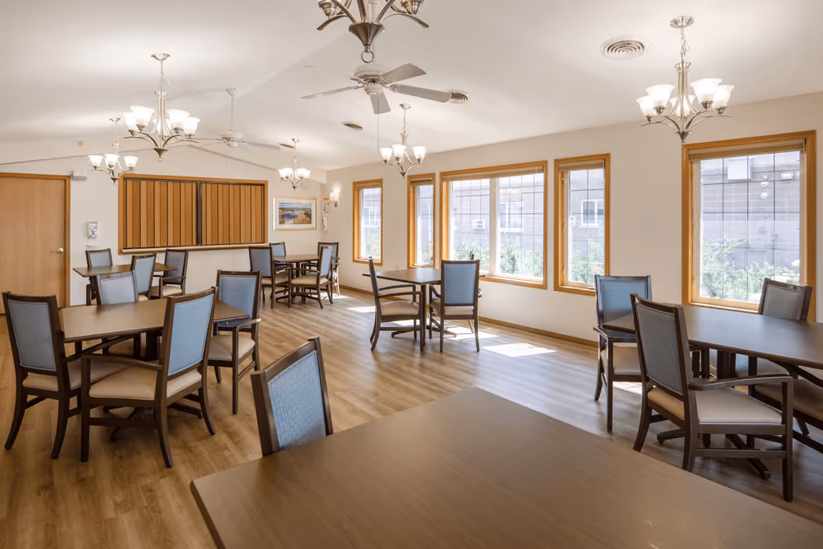 A bright dining room in a senior living facility with multiple wooden tables and chairs arranged neatly. The room has large windows letting in natural light, ceiling fans, and chandeliers providing additional lighting. The floor is wooden, and the walls are light-colored with some framed artwork.