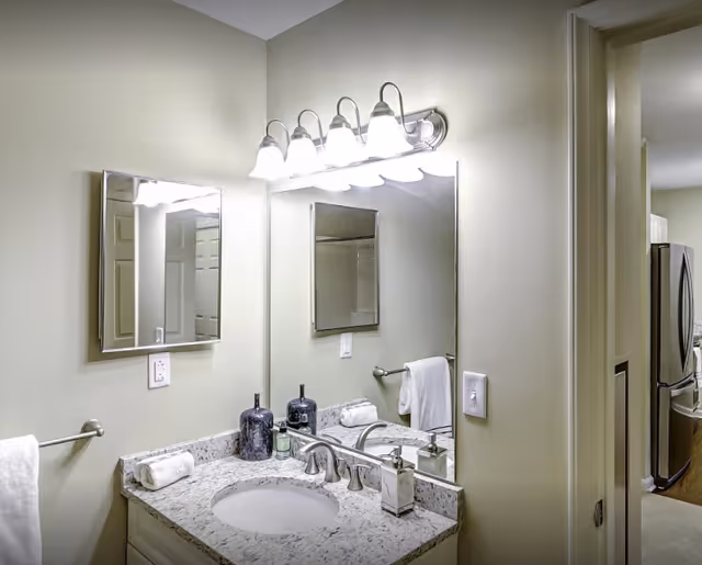 A bathroom vanity area with a granite countertop, an oval sink, and a silver faucet. Above the sink is a large mirror with a light fixture featuring five bulbs. On the countertop are two decorative dark blue bottles, a soap dispenser, and a rolled white towel. A towel rack with a white towel is mounted on the wall to the left. The bathroom walls are painted light beige. To the right, an open doorway leads to a kitchen area with a stainless steel refrigerator visible.