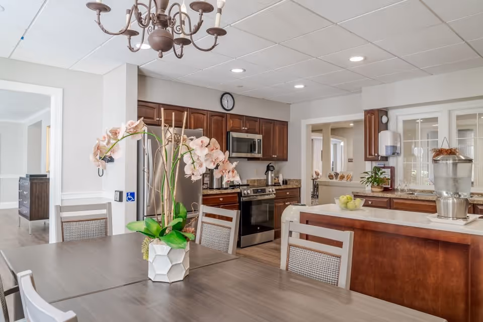A bright and clean kitchen and dining area in a senior living facility. The kitchen features dark wood cabinets, stainless steel appliances including a refrigerator, stove, and microwave. A large island with a water dispenser and a bowl of green apples sits in the center. The dining table in the foreground has a decorative vase with pink orchids. The room has a white ceiling with recessed lighting and a brown chandelier above the table.