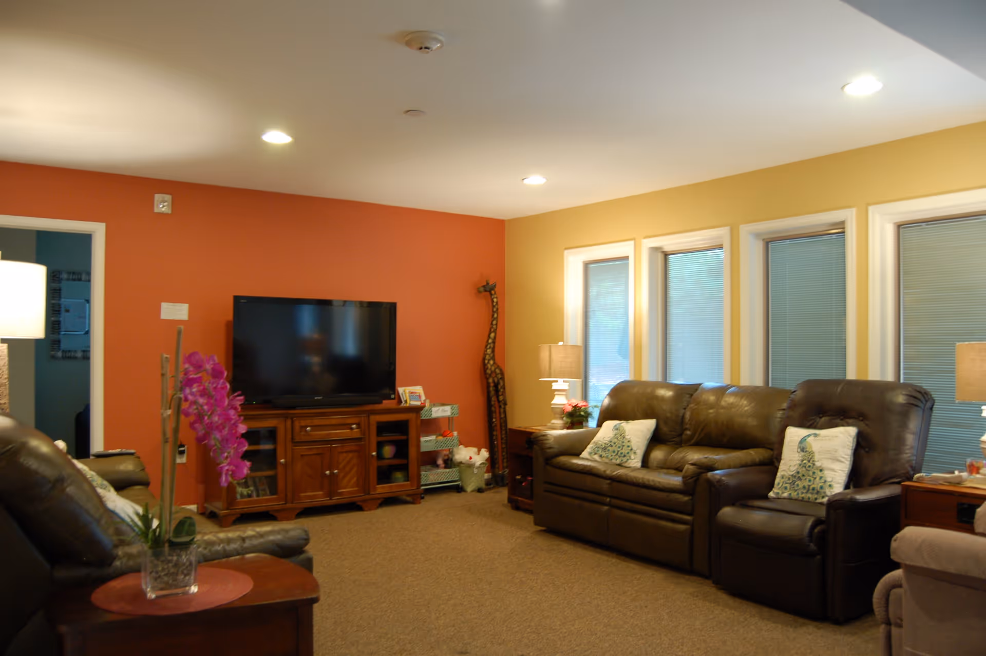 A cozy living room with brown leather sofas and recliners, decorative pillows, a wooden TV stand with a flat-screen television, a tall giraffe sculpture in the corner, table lamps, and a purple orchid on a side table. The walls are painted in warm tones of orange and yellow, and there are three windows with blinds.