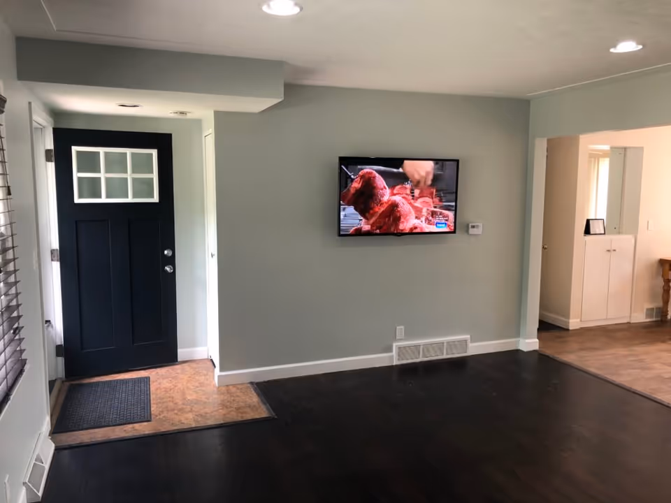 Interior view of a room with a black front door featuring window panes, a wall-mounted flat screen TV displaying a cooking show, dark wood flooring, and light-colored walls. There is an open doorway leading to another room with a cabinet and a mirror.