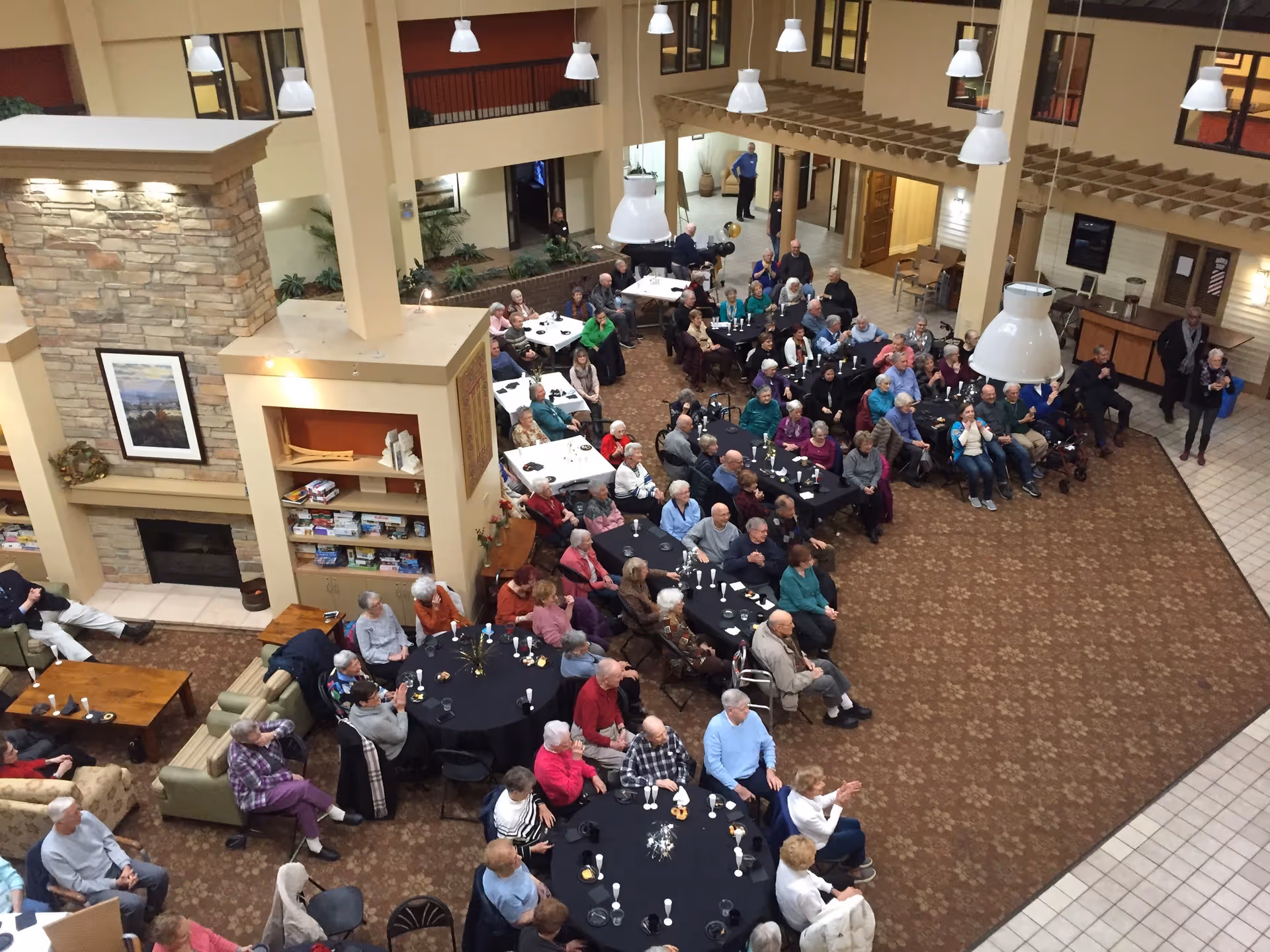 A large group of elderly people seated at round and rectangular tables in a spacious common area with high ceilings, pendant lights, a stone fireplace, and bookshelves. The room has a warm, inviting atmosphere with carpeted floors and some people appear to be engaged in an event or gathering.