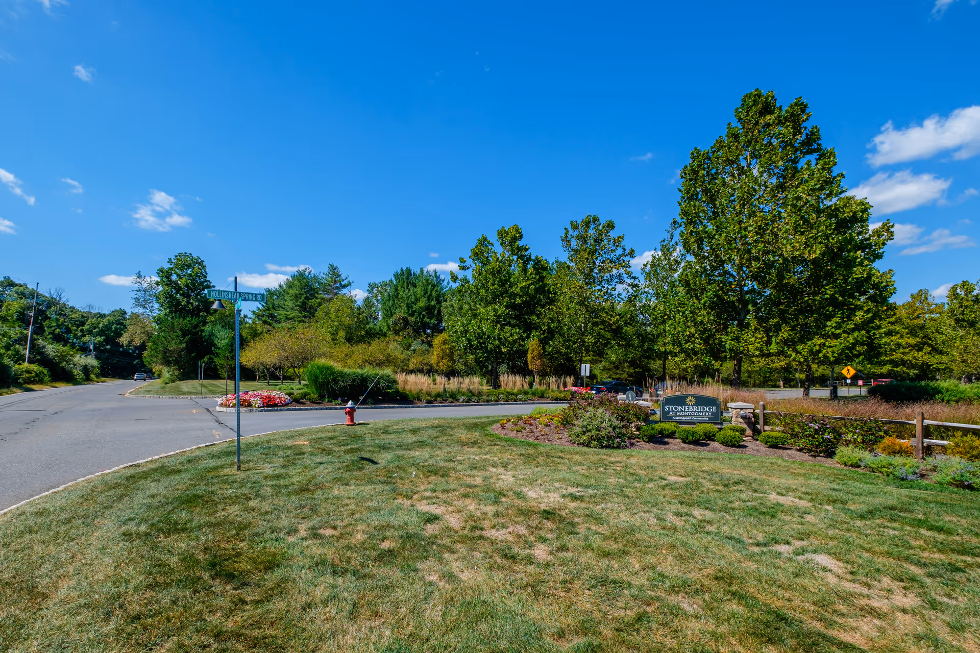 Outdoor view of the entrance area to Stonebridge at Montgomery senior living facility, featuring a grassy lawn, flower beds, trees, a street sign for Hollinshead Spring Rd, and a clear blue sky.