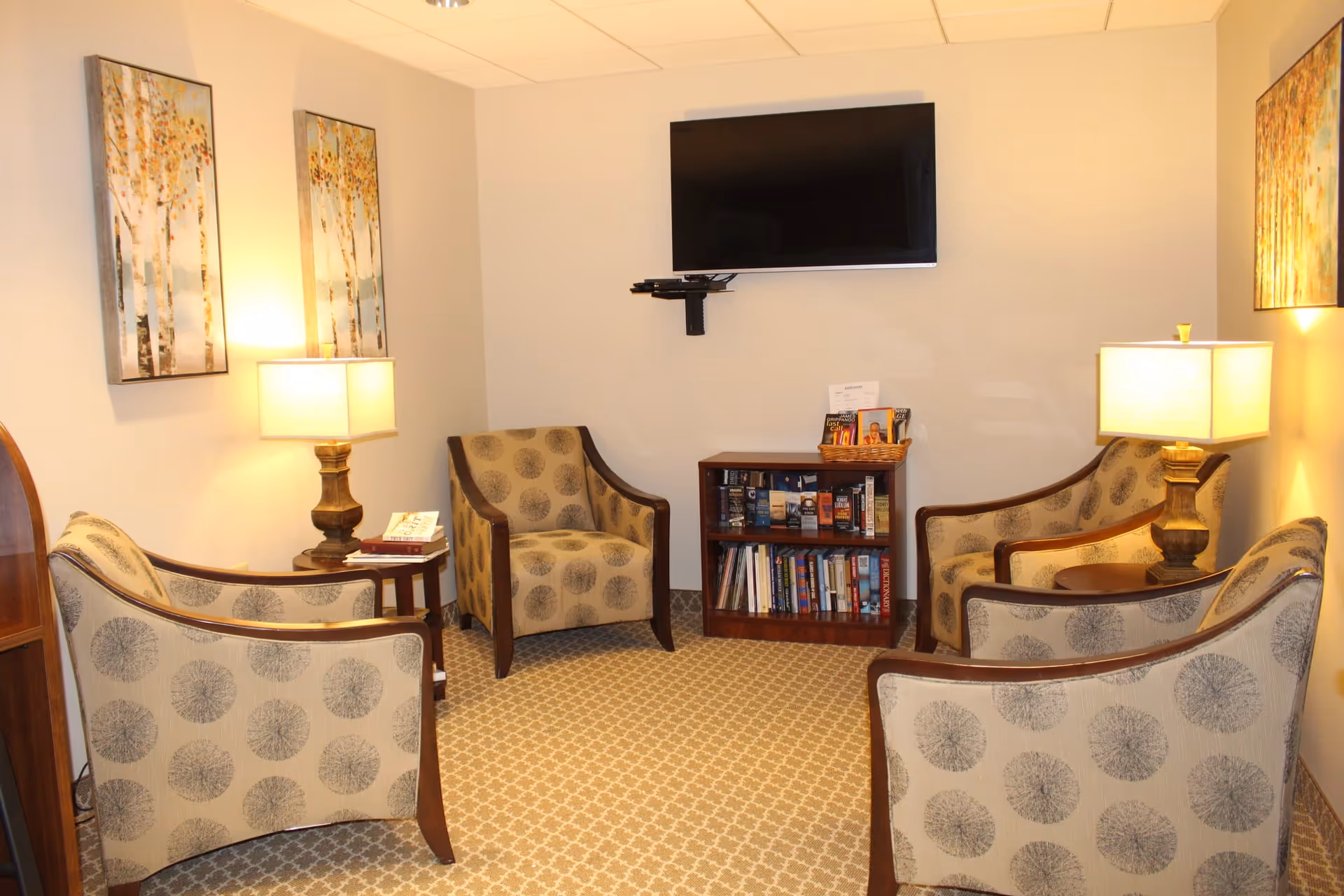 A cozy sitting area in a senior living facility with four patterned armchairs arranged around two wooden side tables with lamps. A small bookshelf filled with books and brochures is against the wall beneath a mounted flat-screen TV. The walls are decorated with framed artwork featuring trees.
