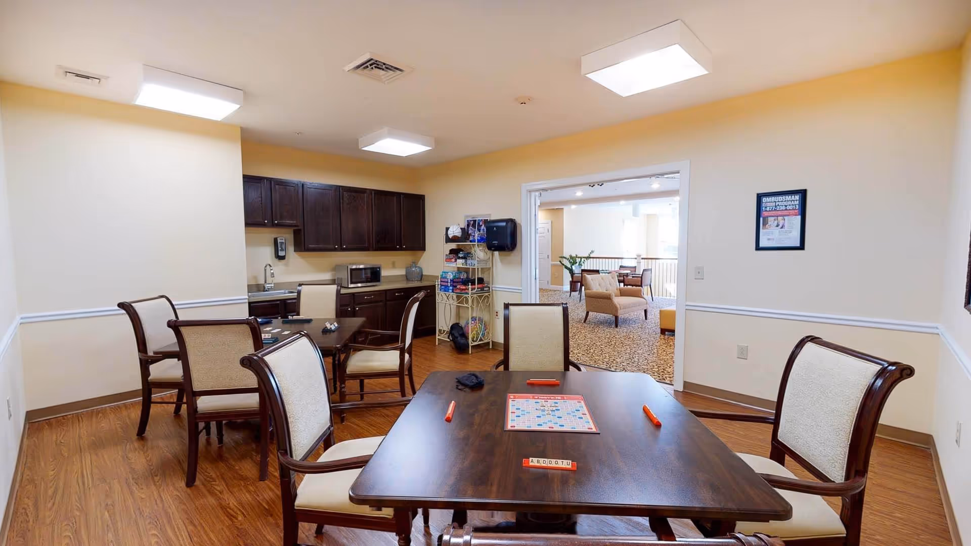 A well-lit common room in a senior living facility featuring a wooden table with a Scrabble game set up, surrounded by cushioned chairs. In the background, there is a kitchenette area with dark wooden cabinets, a microwave, and a sink. Beyond the room, an open doorway leads to a lounge area with additional seating and a carpeted floor.