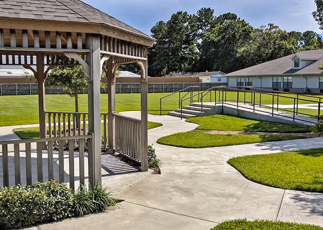 A wooden gazebo with a shingled roof stands on a concrete pathway surrounded by well-maintained green grass and landscaping. In the background, there is a building with a gray roof and white walls, along with a ramp and stairs with handrails leading up to it. Trees and a wooden fence are visible further behind the building under a clear blue sky.