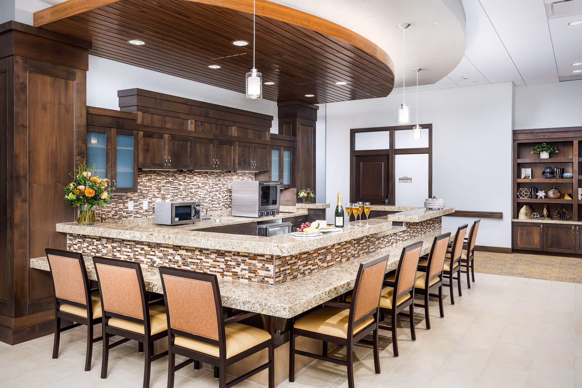 A modern kitchen area with a large granite countertop island surrounded by eight chairs. The kitchen features dark wooden cabinets, a mosaic tile backsplash, and stainless steel appliances including a microwave and oven. There are pendant lights hanging from a wooden ceiling accent above the island. A bottle of champagne and glasses are placed on the counter, with decorative flowers on the side. In the background, there is a wooden door and built-in shelving with decorative items.