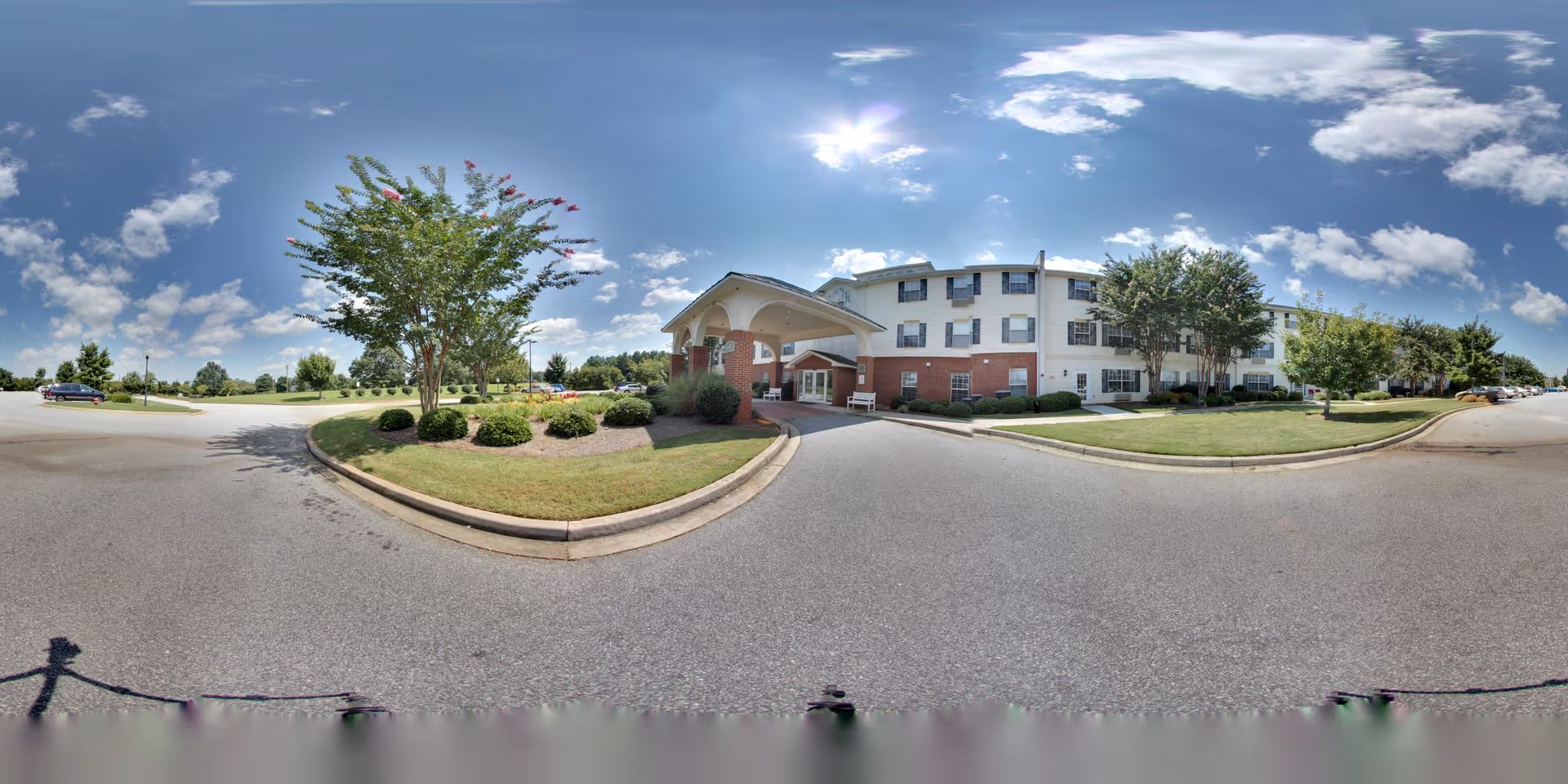 Exterior view of The Legacy of Anderson senior living facility on a sunny day with a clear blue sky and scattered clouds. The building is three stories tall with a brick and white facade, surrounded by well-maintained landscaping including trees, bushes, and grass. A covered entrance is visible with a driveway and parking area in front.