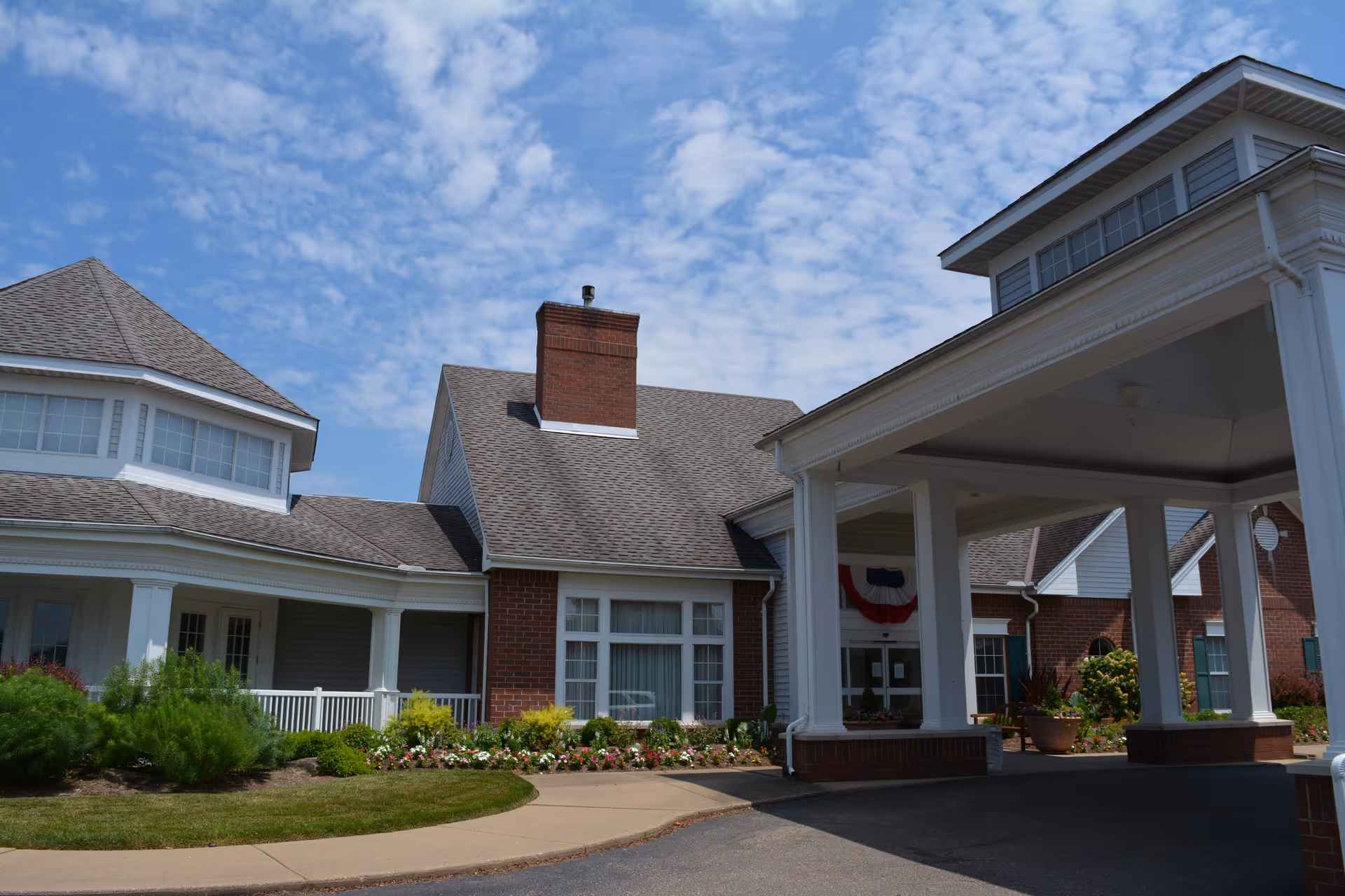Exterior view of The Glenwood Community building with a covered entrance, brick walls, white columns, and a well-maintained garden with flowers and shrubs under a partly cloudy sky.