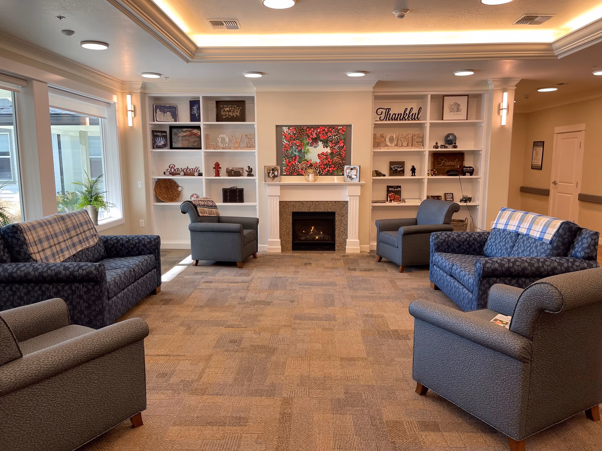 A cozy living room area in a senior living facility with four armchairs and two loveseats arranged around a fireplace. The fireplace is framed by built-in white shelves decorated with various items including framed pictures, decorative words like 'LOVE', 'Thankful', and 'HOME', and small ornaments. Large windows on the left side let in natural light, and the ceiling has recessed lighting with a tray ceiling design.