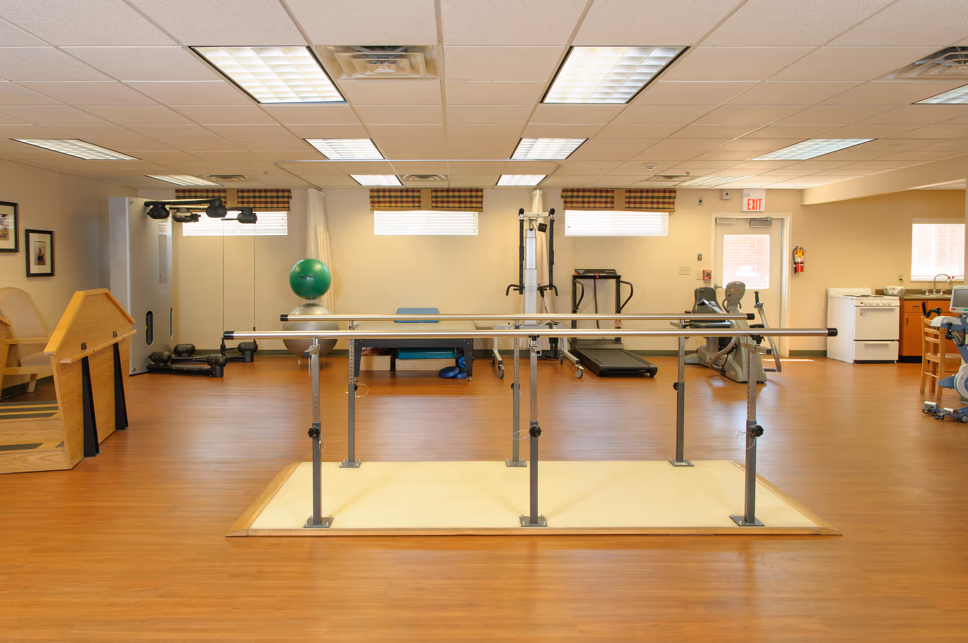 A spacious rehabilitation or physical therapy room with wooden flooring, parallel bars for walking exercises in the center, exercise equipment including a treadmill, stationary bike, exercise balls, and weights along the walls. The room has large windows with plaid valances, fluorescent ceiling lights, and a small kitchenette area with a stove and sink on the right side.