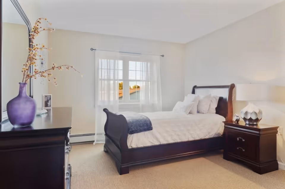 A bright bedroom with a large dark wooden sleigh bed made up with white bedding and multiple pillows. Next to the bed is a matching dark wooden nightstand with a decorative lamp and a small clock. On the left side of the image is a dark wooden dresser with a large mirror above it and a purple vase with dried branches. A window with sheer white curtains allows natural light to fill the room.