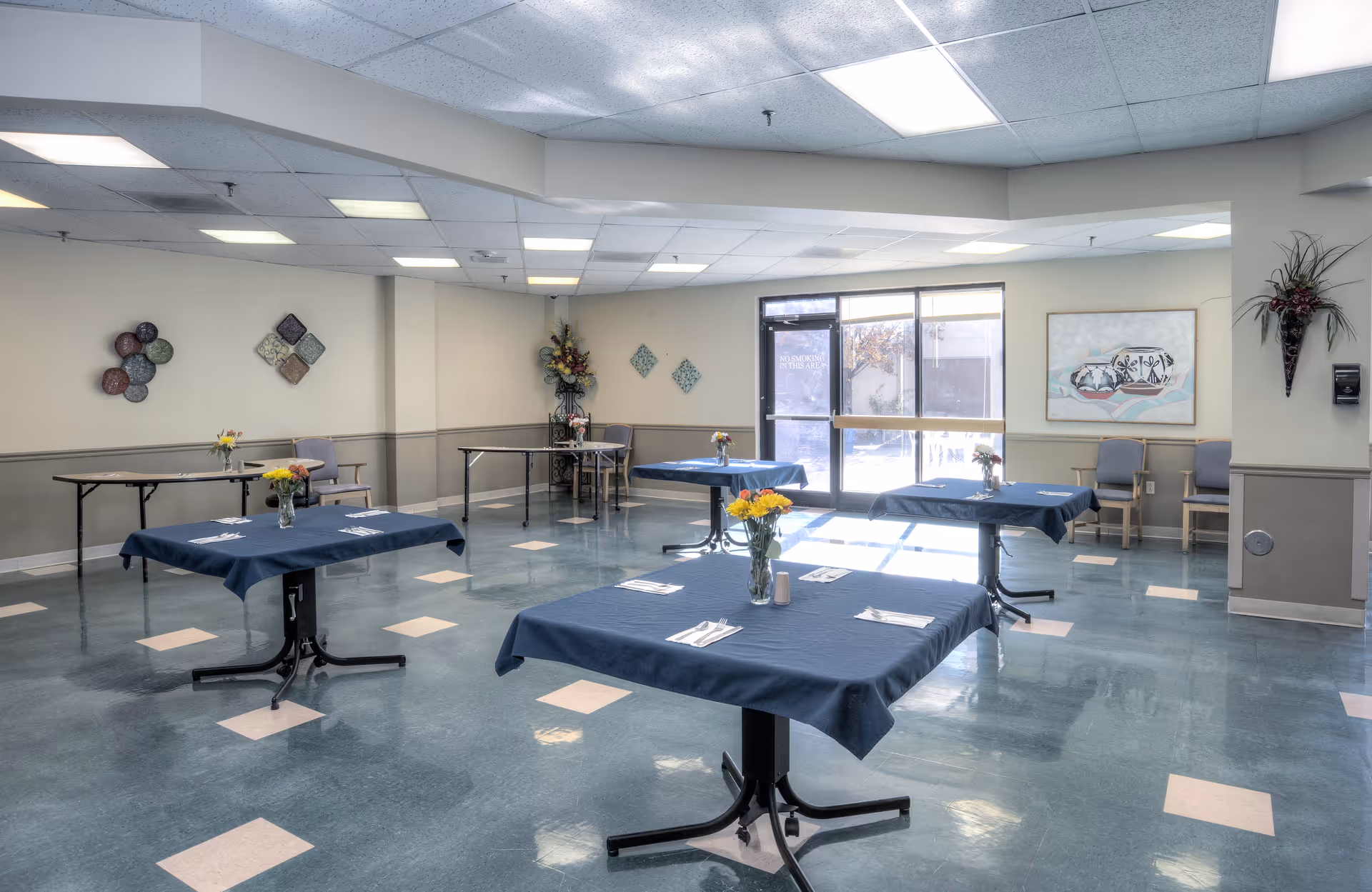 Communal dining room with spaced tables draped in blue tablecloths, chairs, and floral centerpieces under fluorescent ceiling lights.